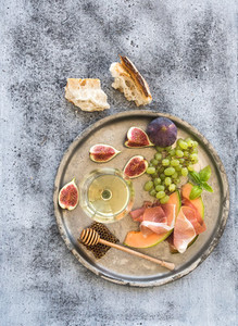 Wine appetizer set Glass of white wine honeycomb with drizzlier figs grapes melon and prosciutto on silver tray over rustic grunge surface Top view