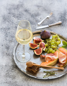 Wine appetizer set Glass of white wine honeycomb with drizzlier figs grapes melon and prosciutto on silver tray over rustic grunge surface