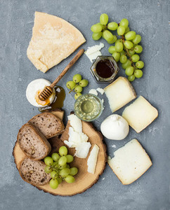 Cheese appetizer selection or wine snack set  Variety of italian cheese  green grapes  bread slices and honey on round wooden board over grey concrete backdrop  top view