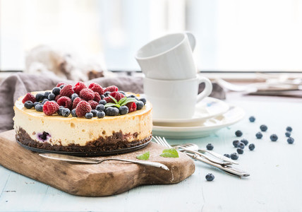 Cheesecake with fresh raspberries and blueberries on a wooden serving board plates cups kitchen napkin silverware over blue background window at the backdrop