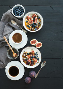 Healthy breakfast set Bowls of oat granola with yogurt fresh blueberries and figs coffee honey over black wooden backdrop
