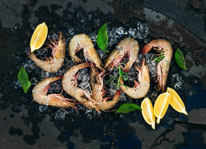Fresh uncooked shrimps with lemon herbs and spices on chipped ice over dark slate stone backdrop horizontal