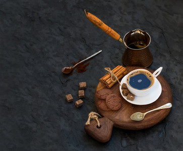 Cup of black coffee with chocolate biscuits  cinnamon sticks and cane sugar cubes on rustic wooden board over dark stone backdrop