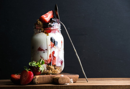 Yogurt oat granola with strawberries  mulberries  honey and mint leaves in tall glass jar on black backdrop