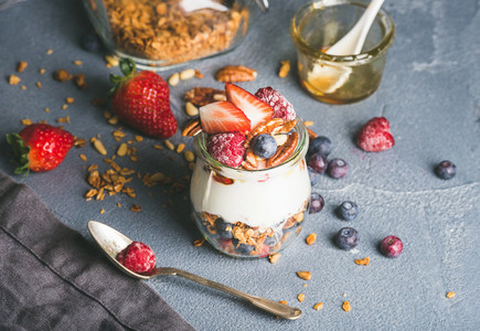 Yogurt oat granola with fresh berries  nuts  honey and mint leaves in glass jar on grey concrete textured backdrop
