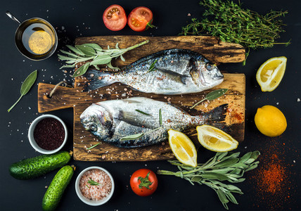 Fresh uncooked dorado or sea bream fish with lemon  herbs  oil  vegetables and spices on rustic wooden board over black backdrop