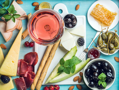 Summer wine snack set  Glass of rose  meat  cheese  olives  honey  bread sticks  nuts  capers and berries with white ceramic board in center  blue wooden background