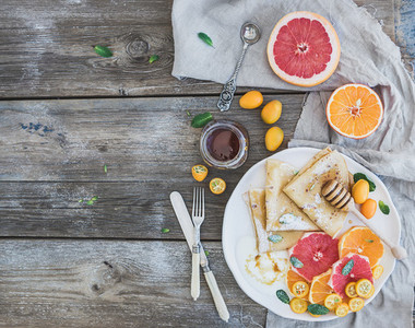 Spring vitamin breakfast set  Thin crepes or pancakes with fresh grapefruit  orange  kumquat  honey  cream and mint leaves over a rustic wood background