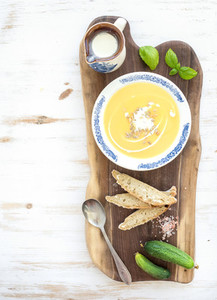 Pumpkin soup with cream  fresh basil  cucumbers and bread in vintage ceramic plate on wooden board over white background  top view
