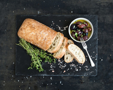 Italian ciabatta bread cut in slices on wooden chopping board with herbs  garlic and olives over dark grunge backdrop  copy space