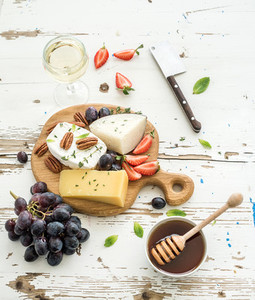Cheese appetizer selection or wine snack set  Variety of cheese  grapes  pecan nuts  strawberry and honey on round wooden board over rustic white backdrop