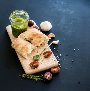Pesto sauce in jar ciabatta bread cherry tometoes thyme and garlic on rustic wooden board over black grunge backdrop