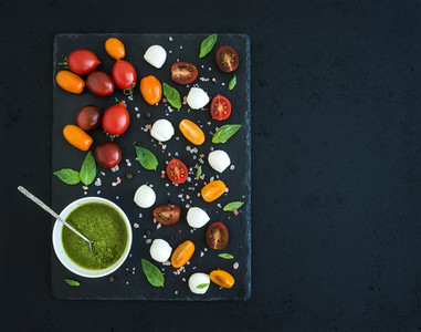 Caprese set Cherry tomatoes of various color mozarella basil leaves spices and pesto sauce on black slate tray over dark grunge background Top view