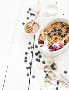 Healthy breakfast Oat granola berry crumble with fresh blueberries yogurt and honey in ceramic baking dish over white rustic backdrop