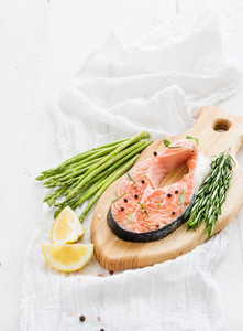 Raw salmon steak with asparagus  lemon  spices and rosemary on rustic wooden chopping board over white backdrop