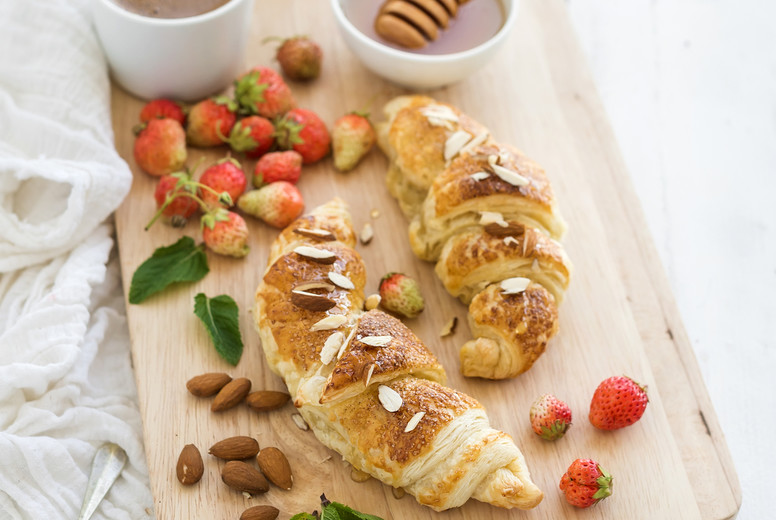 Freshly baked almond croissants with garden strawberries, mint, honey and coffee on serving board over white rustic wood backdrop