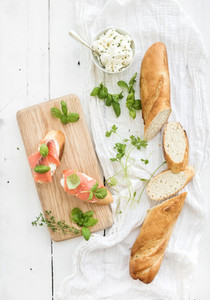 Salmon ricotta and basil sandwiches with baguette on a rustic wooden board over white wood background Top view