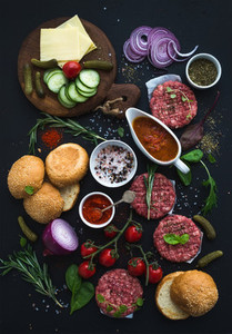 Ingredients for cooking burgers  Raw ground beef meat cutlets  buns  red onion  cherry tomatoes  greens  pickles  tomato sauce  cheese  herbs and spices over black background  top view