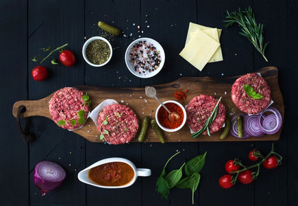 Ingredients for cooking burgers Raw ground beef meat cutlets on wooden chopping board red onion cherry tomatoes greens pickles tomato sauce cheese herbs and spices over black background top v