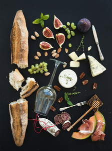Wine and snack set  Baguette  glass of white  figs  grapes  nuts  cheese variety  meat appetizers  herbs on black grunge background  top view