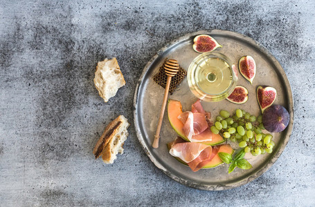 Wine appetizer set  Glass of white wine  honeycomb with drizzlier  figs  grapes  melon and prosciutto on silver tray over rustic grunge surface  Top view
