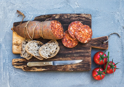 Wine snack set Hungarian mangalica pork salami sausage rustic bread and fresh tomatoes on dark wooden board over a rough grey blue concrete background