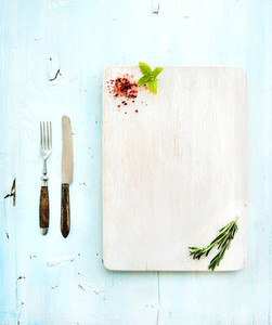 Kitchen ware set  White wooden chopping board  knife  fork  spices and herbs on a light blue background