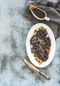Chocolate Belgian waffles with salted caramel sauce and mint on white ceramic serving plate over grunge background  top view