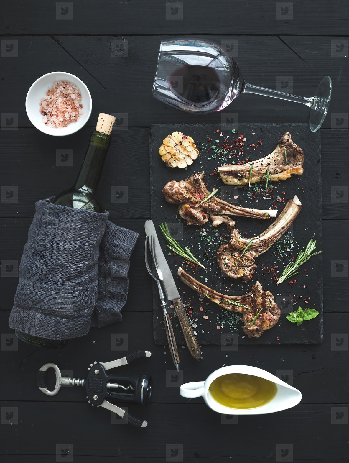 Grilled lamb chops. Rack of Lamb with garlic, rosemary, spices on slate tray, wine glass, oil in a saucer and bottle over black wood background