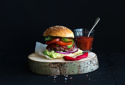 Fresh homemade burger on wooden serving board with spicy tomato sauce sea salt and herbs over black  background