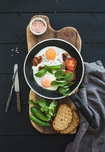Pan of fried eggs  bacon  tomatoes with bread  mangold and cucumbers on rustic wooden serving board over dark table surface