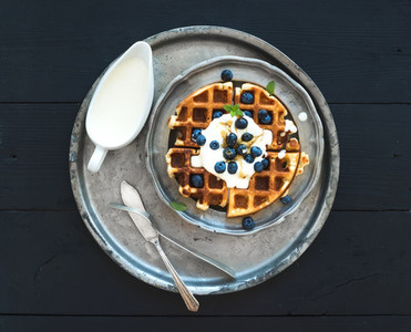 Soft Belgian waffles with blueberries  honey and whipped cream on vintage metal plate over black wooden background  top view