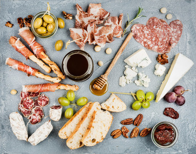 Cheese and meat appetizer selection or wine snack set Variety of  cheese salami prosciutto bread sticks baguette honey grapes olives sun dried tomatoes pecan nuts over grey concrete textured