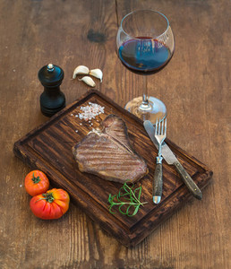 Cooked meat t bone steak on serving board with garlic cloves  tomatoes  rosemary  spices and glass of red wine over rustic wooden background