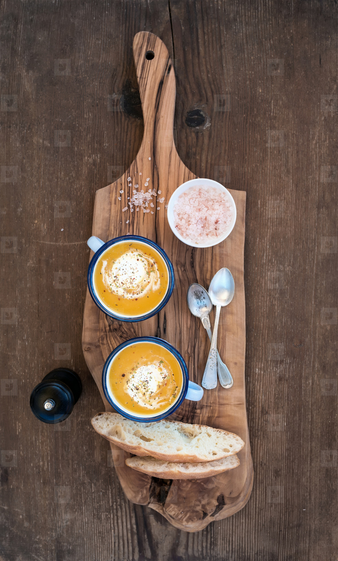 Homemade pumpkin cream soup in enamel mugs with herbs and fresh bread slices on olive serving board over rustic wooden background
