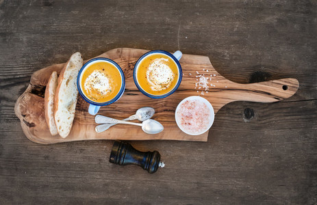 Homemade pumpkin cream soup in enamel mugs with herbs and fresh bread slices on olive serving board over rustic wooden background  horizontal