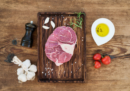 Raw fresh beef meat cross cut for ossobuco with garlic cloves  cherry tomatoes  rosemary  pepper  oil and salt on serving board over rustic wooden background
