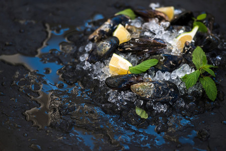 Fresh uncooked mussels with lemon herbs and spices on chipped ice over dark slate stone backdrop