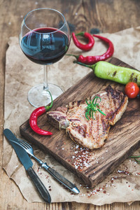 Cooked meat t bone steak on serving board with roasted tomatoes  chili peppers  fresh rosemary  spices and glass of red wine over rustic wooden background