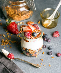 Yogurt oat granola with fresh berries  nuts  honey and mint leaves in glass jar on grey concrete textured backdrop