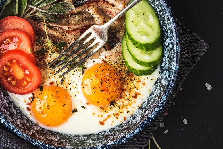 Breakfast set. Pan of fried eggs with bacon, fresh tomato, cucumber, sage and bread on dark serving board over black background