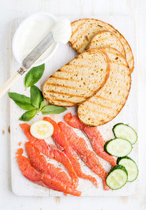 Ingredients for healthy sandwich  Grilled bread slices  smoked salmon  cottage cheese  cucumber nd basil on white wooden board