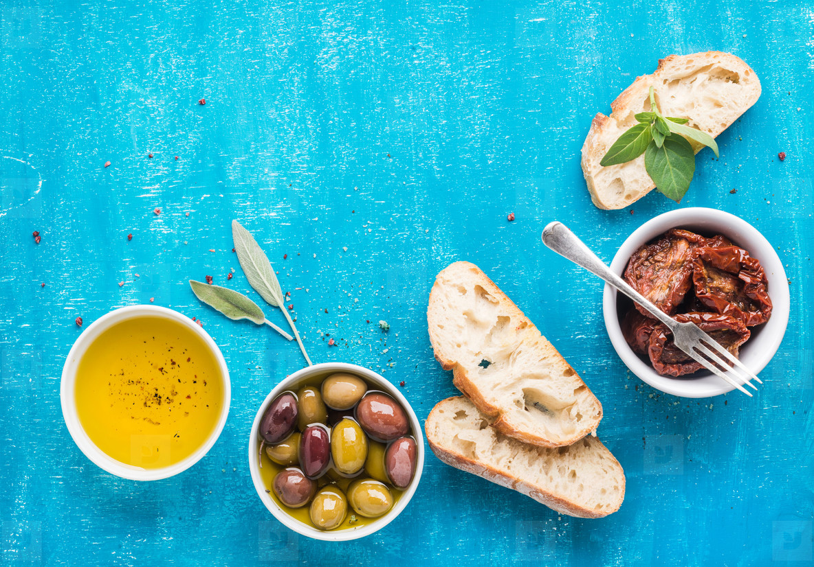 Mediterranean snacks set. Olives, oil, sun-dried tomatoes, herbs and sliced ciabatta bread on over blue painted background