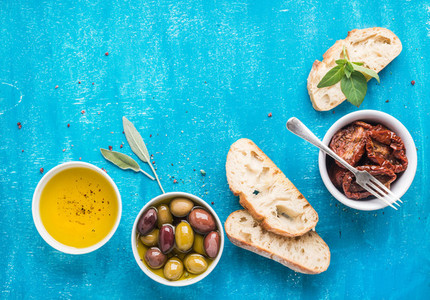 Mediterranean snacks set Olives oil sun dried tomatoes herbs and sliced ciabatta bread on over blue painted background