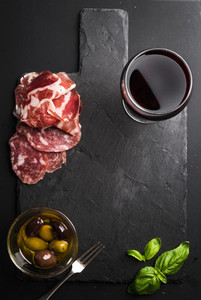 Glass of red wine meat appetizer olives and basil on black  slate stone board over dark background