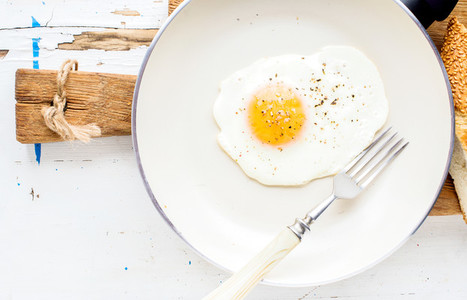 Fried egg with spice and bread slices in white ceramic frying pan on wooden board