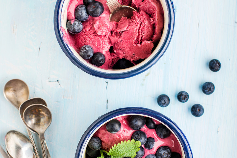 Homemade blueberry ice cream scoops with fresh berries and mint leaves in enamel mugs over light blue background