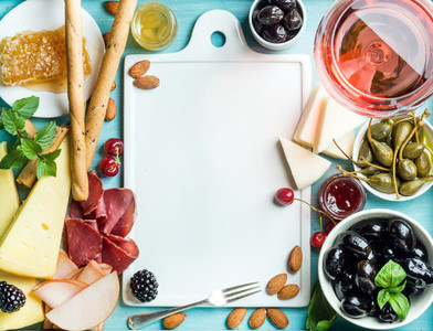 Summer wine snack set Glass of rose meat cheese olives honey bread sticks nuts capers and berries with white ceramic board in center blue wooden background