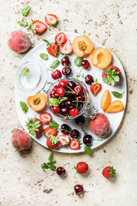 Healthy summer fruit variety Sweet cherries strawberries peaches apricots and mint leaves on white ceramic serving plate over light concrete background