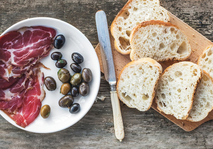 Smoked meat or prosciutto and olives on a white plate vintage knife baguette slices over rough wood background